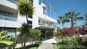 Un hotel de diseño moderno, con una entrada central y un paseo techado, rodeado de palmeras y otra vegetación.