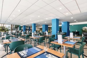 A spacious cafeteria with tables and chairs, a high ceiling, and blue pillars. - Catalonia Oro Negro (Playa de las Americas)