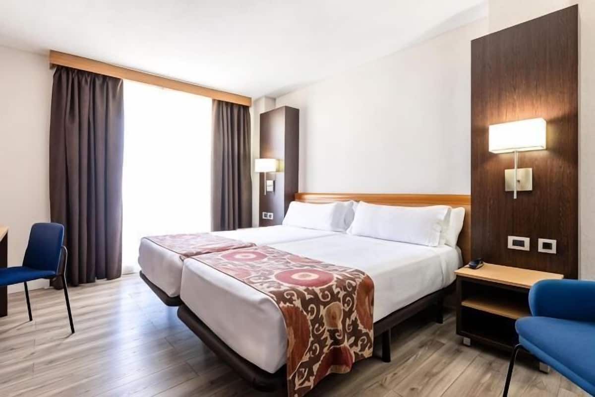 Ein Hotelzimmer mit Doppelbett, zwei Stühlen, einem Schreibtisch und einem großen Fenster mit Vorhängen.