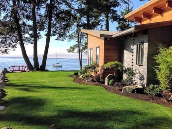 A modern house with a well-maintained lawn, a wooden deck, and a view of the ocean. - Secluded, peaceful, ocean front home. Close to all amenities/outdoor activities. (Sooke)