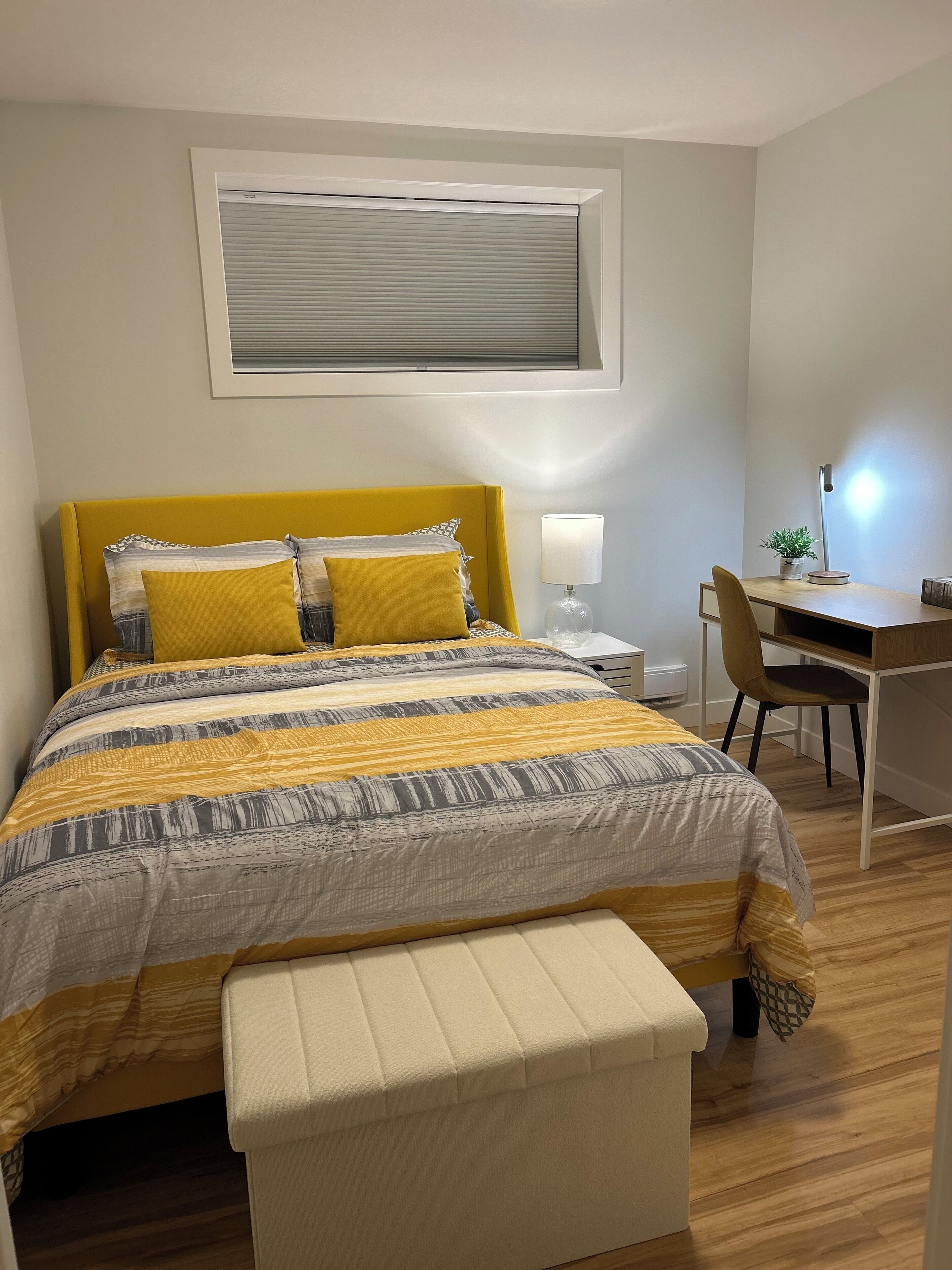 A bedroom with a bed, a desk, a chair, a lamp, and a window with blinds.