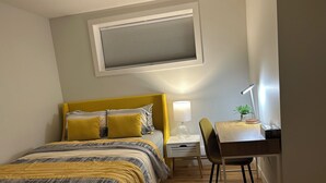 A bedroom with a bed, bedside table, lamp, chair, and a desk with a plant. - The Prairie Nest (Saskatoon)
