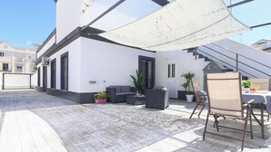 Una terraza en la azotea con mobiliario exterior, una escalera y un edificio blanco al fondo.