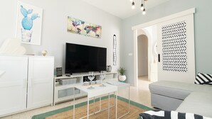 Una sala moderna con televisor de pantalla plana, muebles blancos, un tapete estampado y obras de arte en las paredes.