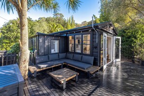 Un espacio exterior moderno con un sofá empotrado y mesas de madera, rodeado por una terraza de madera y abundante vegetación.
