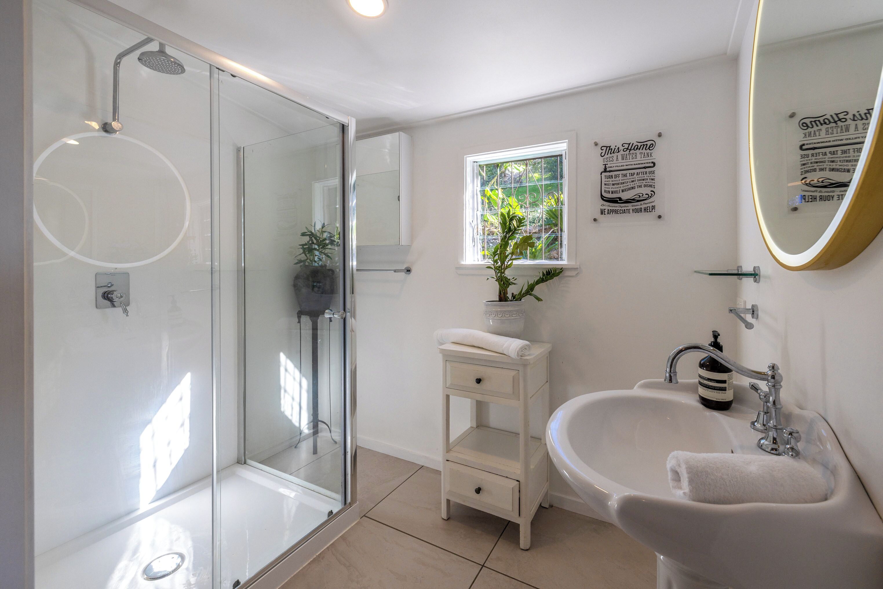 Une salle de bain moderne avec une douche à parois de verre, un lavabo blanc avec une serviette et une fenêtre agrémentée d’une plante.