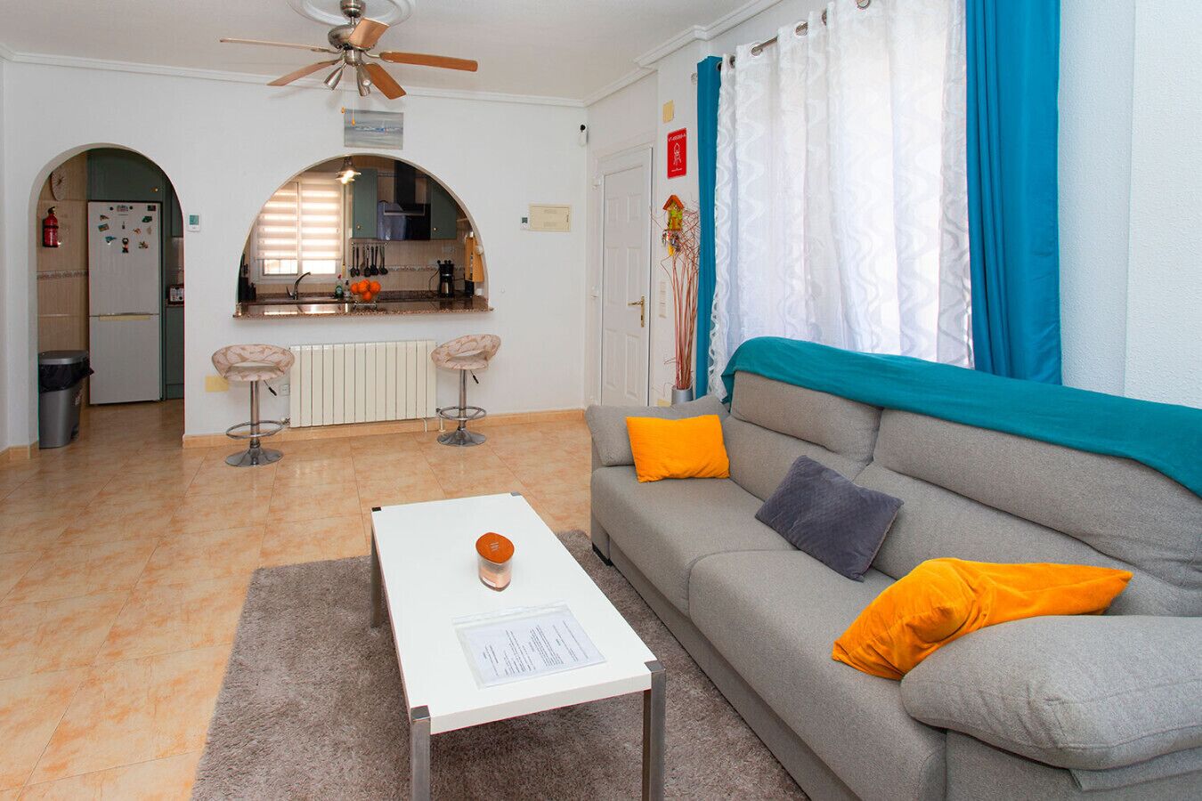 Un salon avec un canapé gris, des coussins orange, une table basse blanche et un ventilateur de plafond.