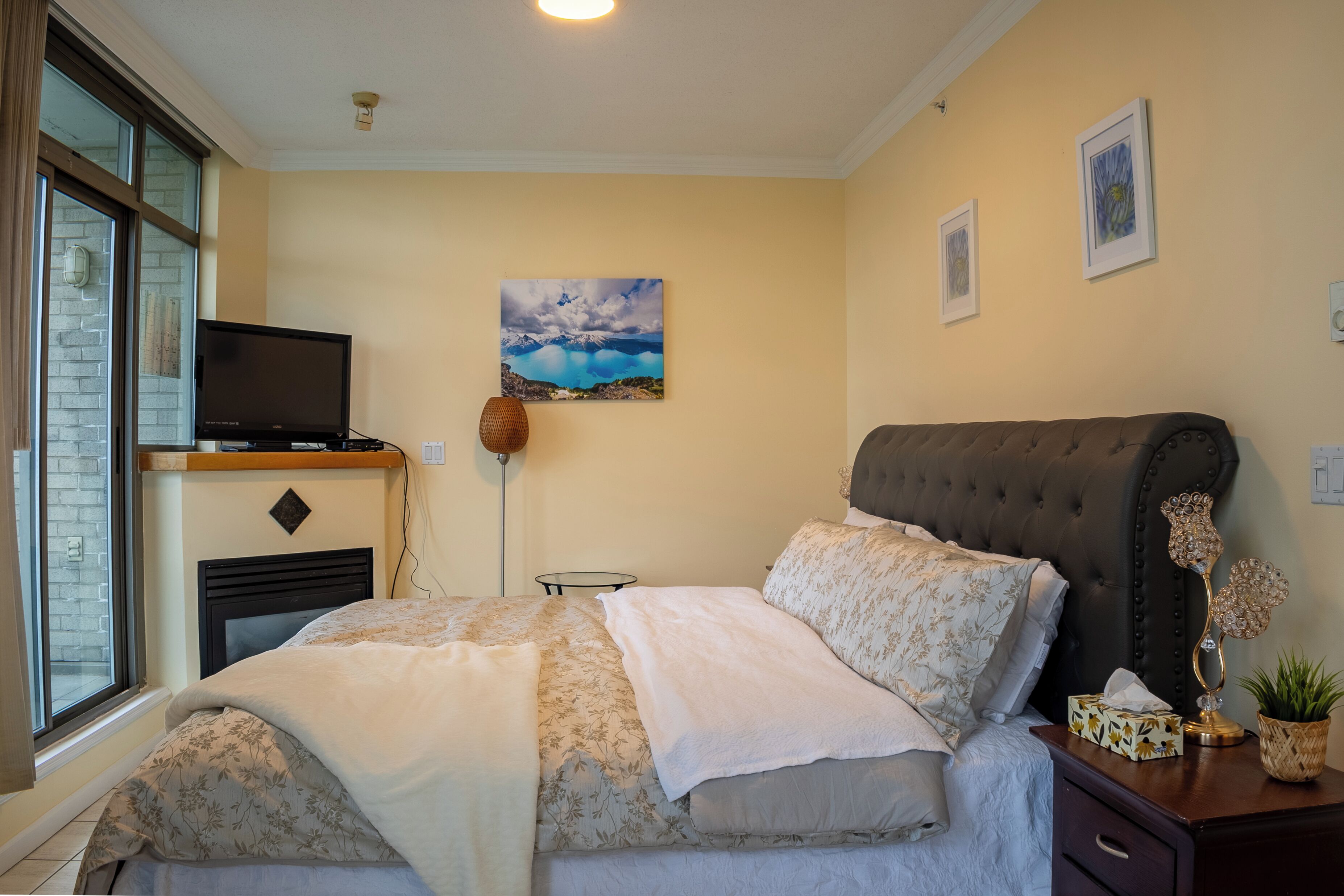 A bedroom with a bed, a television on a stand, a painting on the wall, and a window with a view.