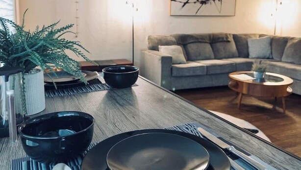 Un comedor con una mesa de madera dispuesta para cuatro personas, un sofá gris y una mesa de centro al fondo.