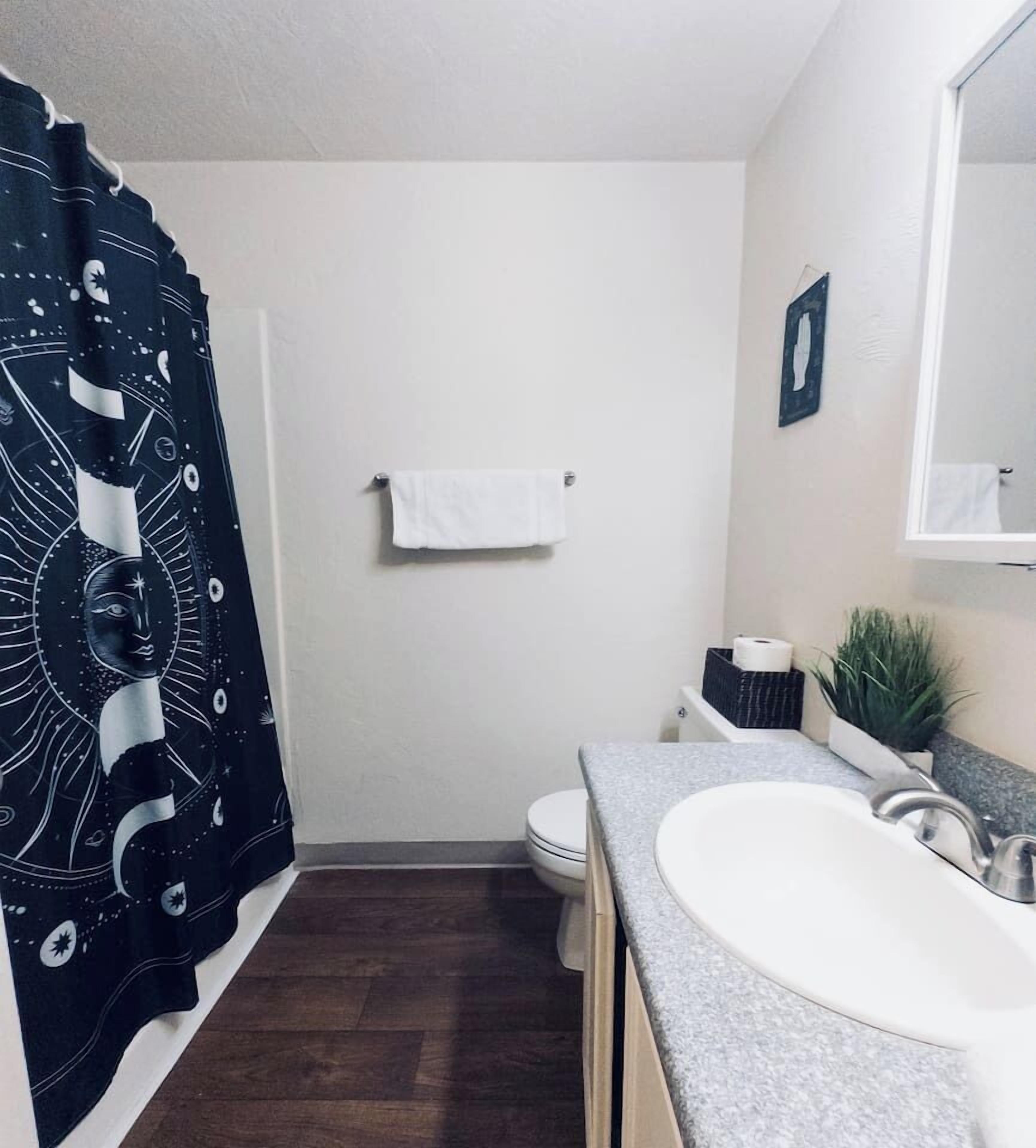 Un baño con un lavabo blanco, inodoro, cortina de ducha con diseño espacial, toalla blanca colgada en la pared y una pequeña planta en maceta sobre la encimera.