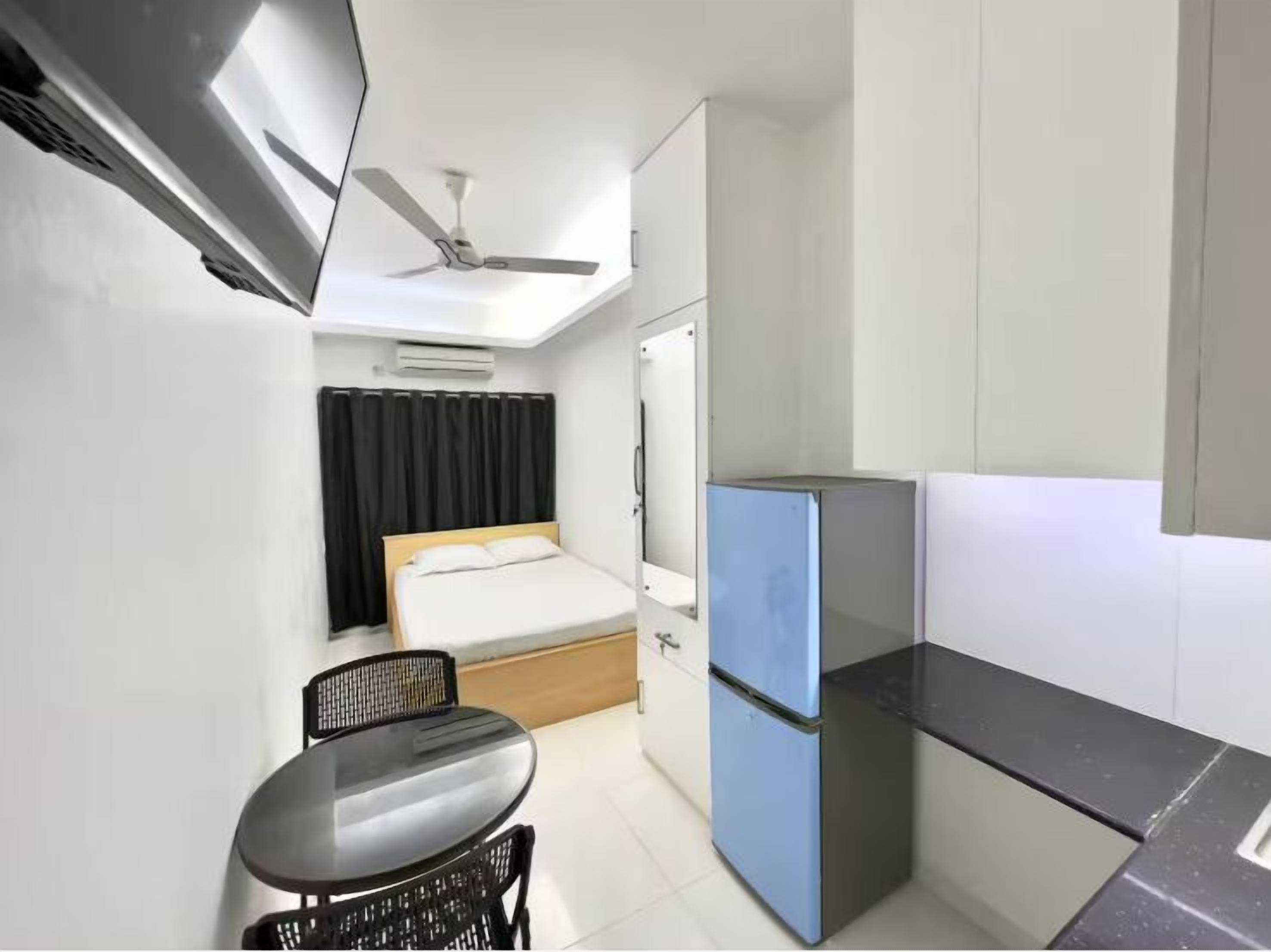 A compact room with a bed, a small table, a chair, a blue refrigerator, and a ceiling fan.