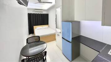 A compact room with a bed, a small table, a chair, a blue refrigerator, and a ceiling fan.