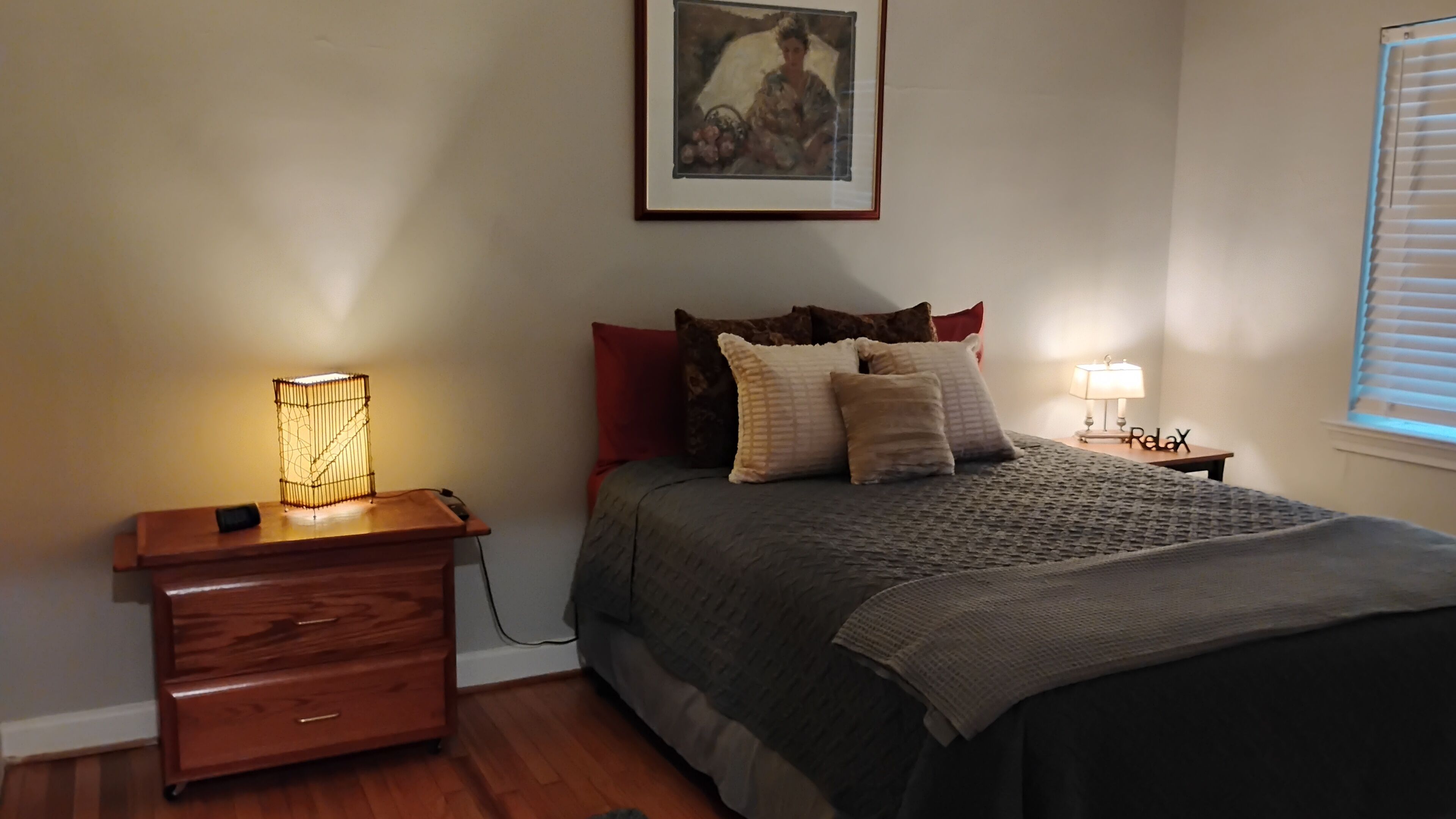 Een slaapkamer met een bed, een nachtkastje met een lamp, een houten dressoir en een schilderij aan de muur.