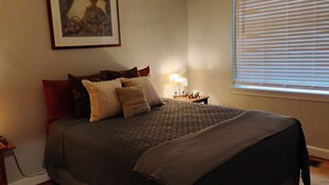 Ein ordentlich gemachtes Bett mit dunkler Decke und mehreren Kissen, ein Nachttisch mit Lampe sowie ein Fenster mit Jalousien.