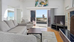 A modern living room with a large sofa, a coffee table, a flat-screen TV, and a fireplace. There is a large window with a view of the beach and a balcony.