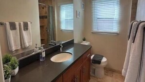 Un baño con encimera oscura, un lavamanos, un espejo y un inodoro. Hay toallas colgadas en la pared y una cortina de ducha.