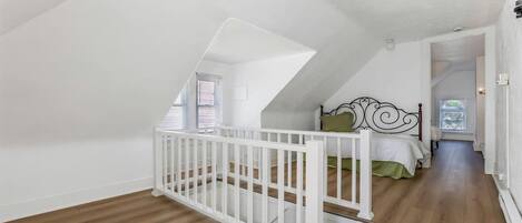 A room with a sloped ceiling, a white crib, a bed with green bedding, and a wooden floor.
