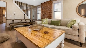 A modern living room with a wooden coffee table, a beige sofa with green cushions, a brick wall, and a staircase. - Franklin Park Gem - Historic meets Modern w/ Space for the Whole Crew - 4BR, 2BA (Columbus)