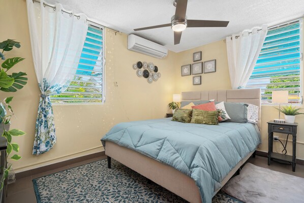 A bedroom with a bed, ceiling fan, air conditioning unit, and a window with curtains.