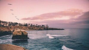 A coastal scene with a rocky shoreline, waves crashing onto the beach, and a row of buildings on a hillside in the background.