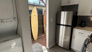 A compact kitchen with white cabinets, a stainless steel refrigerator, and a surfboard leaning against the wall.