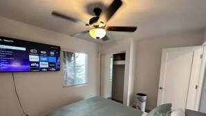 A bedroom with a bed, a ceiling fan, a TV mounted on the wall, and a window with blinds.