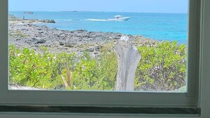 Janela com vista para um litoral rochoso, um barco na água e um tronco de árvore em primeiro plano.