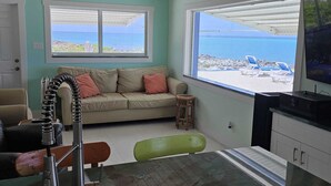 Quarto com sofá, um banquinho pequeno, mesa e televisão. Há duas janelas com vista para a praia e o oceano.