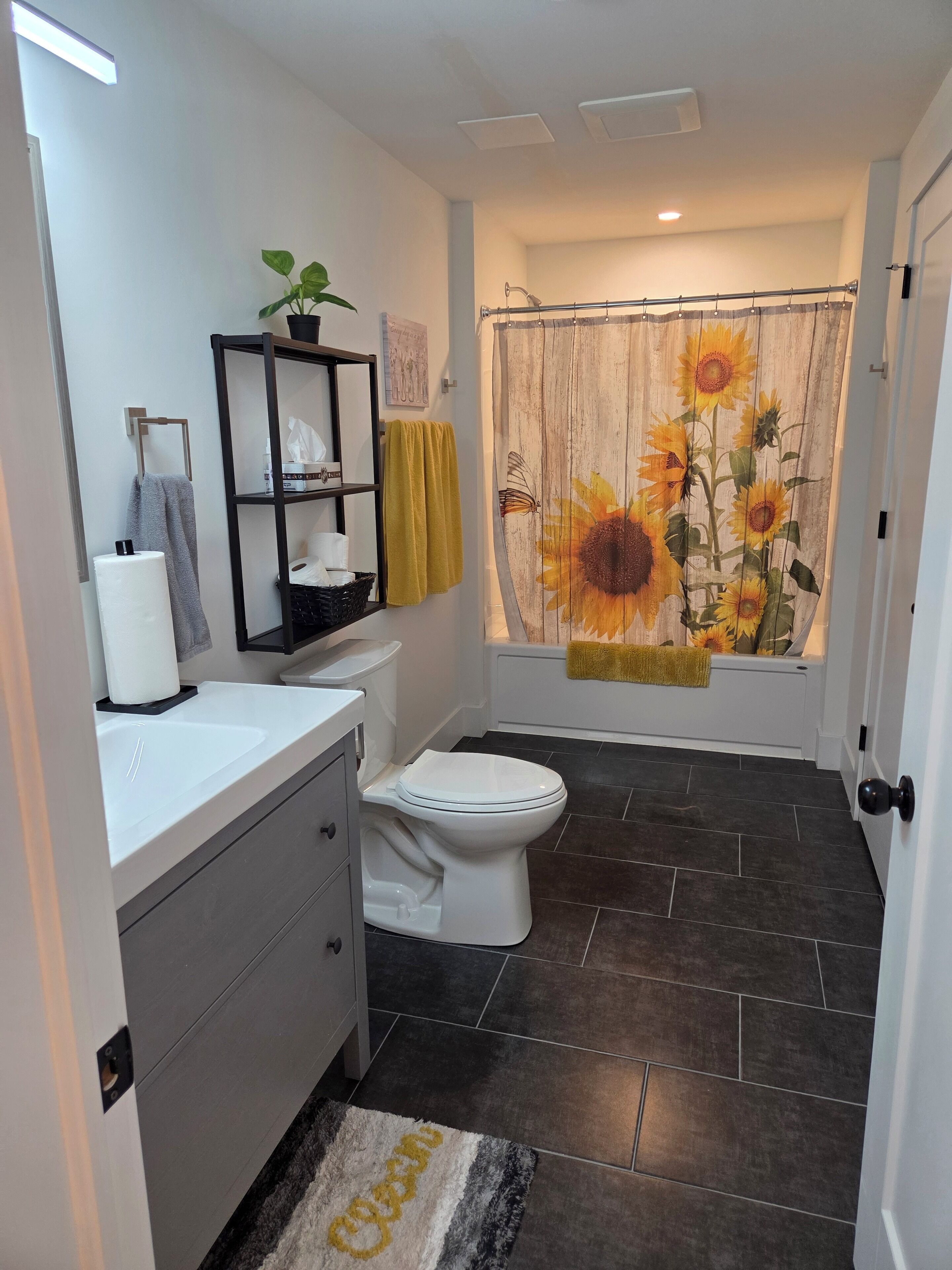 A bathroom with a white toilet, a black shelf with toiletries, a towel rack with a yellow towel, a shower curtain with sunflowers, and a dark tiled floor.