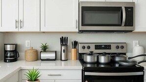 Una cocina moderna con armarios blancos, un microondas de acero inoxidable, una cafetera, un tostador, una estufa con ollas y una pequeña planta.