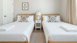 Habitación de hotel con dos camas, una mesita de noche con lámpara, una planta enmarcada en la pared y una puerta.