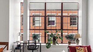 Une chambre avec une grande fenêtre, un bureau, une chaise, un lit et deux plantes en pot.