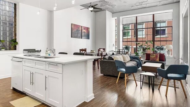 Une cuisine moderne avec des meubles blancs, un îlot central et un coin repas agrémenté de chaises bleues et d’une table en bois.