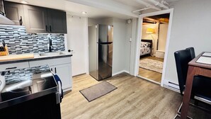 A compact kitchen with modern appliances, a dining area with a wooden table, and a visible bedroom with a bed and lamp. - Your Clean Clubhouse in St Boniface. Perfect for Students/Short term home away! (Winnipeg)