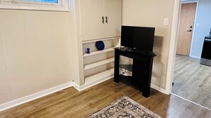 A room with a wooden floor, a television on a black stand, a white cabinet with a blue decorative plate, and a patterned rug. - Your Clean Clubhouse in St Boniface. Perfect for Students/Short term home away! (Winnipeg)