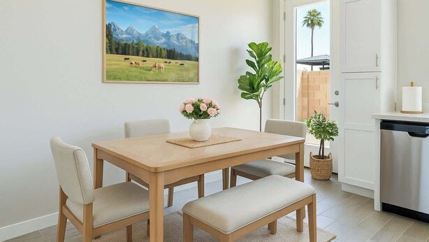 A dining area with a wooden table, four chairs, a vase of flowers, and a painting of a mountain landscape on the wall.