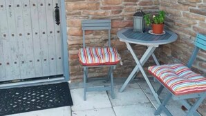 Un porche aux murs de pierre, avec une porte blanche, une petite table sur laquelle est posée une plante en pot, et deux chaises à rayures.