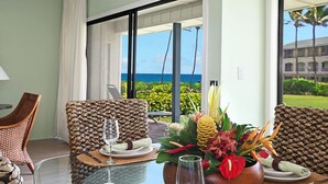 A dining area with wicker chairs, a glass table, and a view of the ocean. - 1st Floor 2BR Ocean View • Steps to Beach • Pool • AC • Sleeps 6 (Koloa)