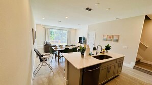 A modern kitchen with a central island, a dining area with a table and chairs, and a view of a garden through large windows. - Beautiful Condo at Reunion (Kissimmee)