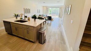 A modern kitchen with a central island, a dining area with a table and chairs, and a view of the outdoors through large windows. - Beautiful Condo at Reunion (Kissimmee)