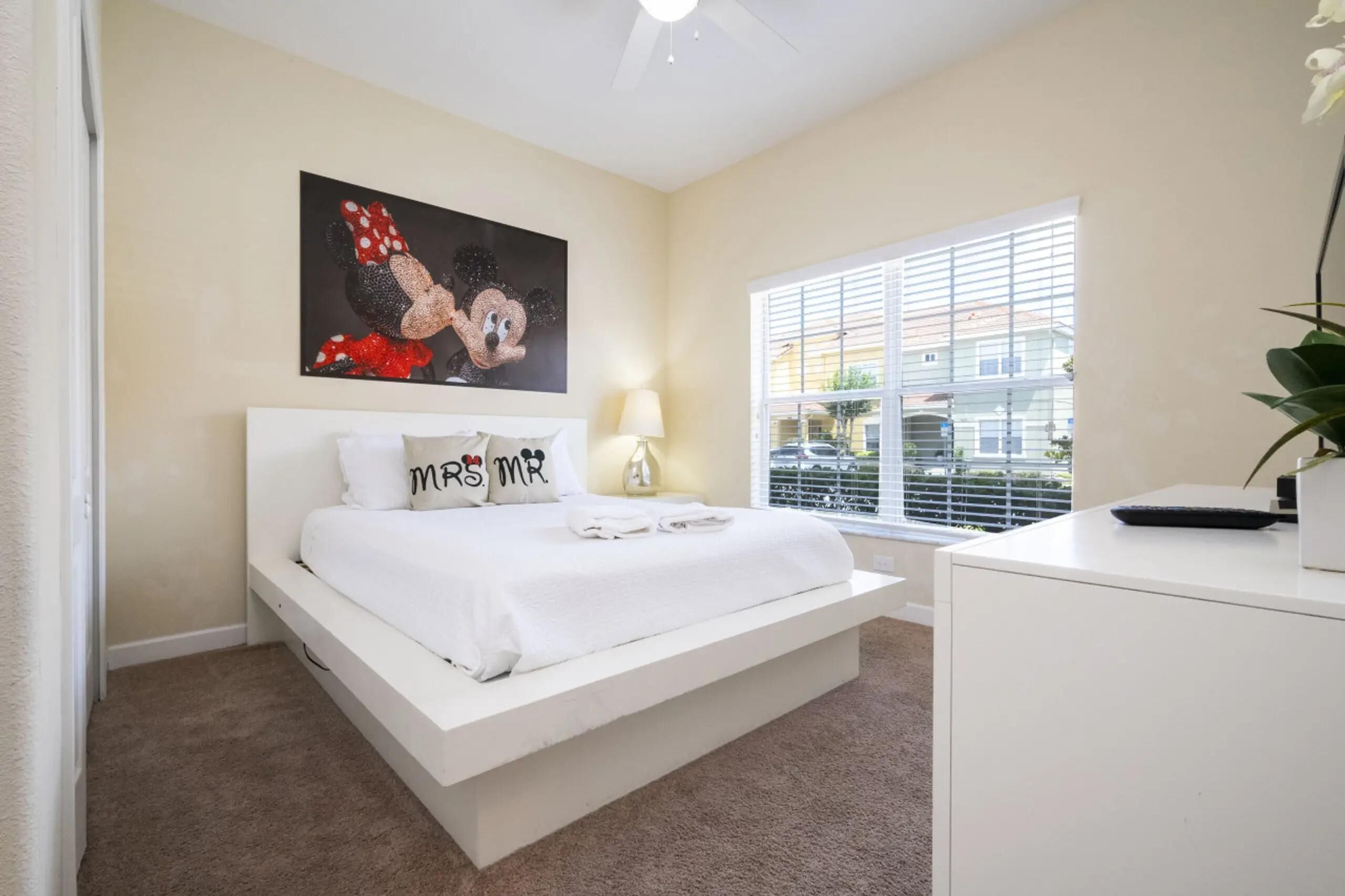 A bedroom with a bed, a desk, a window with blinds, and a ceiling fan.