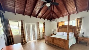 Une chambre spacieuse dotée d’un grand lit à baldaquin en bois, d’un ventilateur de plafond et offrant une vue sur l’extérieur grâce à de grandes fenêtres.