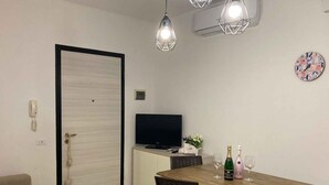 A compact living space with a wooden dining table, two wicker chairs, a TV on a white cabinet, and a door with a heart-shaped handle. - Holiday apartment with air conditioning and washing machine (Rosolina Mare)