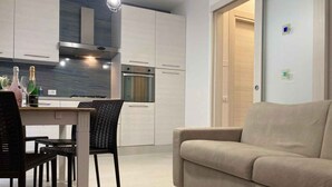 A modern kitchen with white cabinets, a dining area with a table and chairs, and a beige sofa in a living area. - Holiday apartment with air conditioning and washing machine (Rosolina Mare)