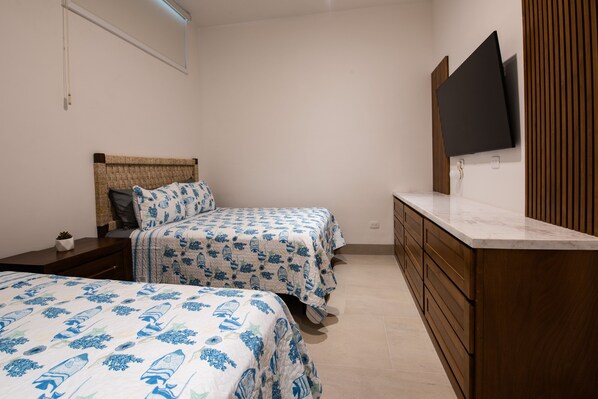 A bedroom with two beds, a TV mounted on the wall, wooden cabinets, and a bedside table with a lamp.