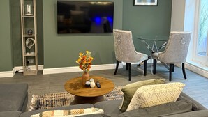 En moderne stue med en grå sofa, et rundt sofabord med en vase blomster, to grå lænestole, et fladskærms-tv og en reol.