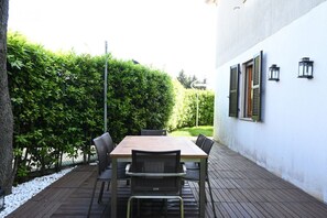 An outdoor patio with a wooden table and chairs, surrounded by a white wall and a hedge.