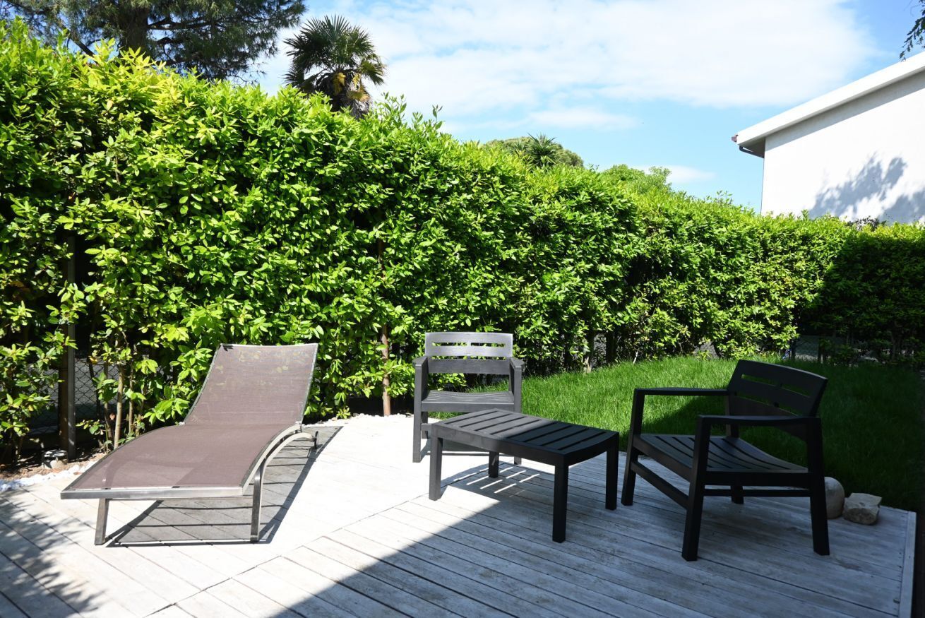 Um terraço com duas espreguiçadeiras e uma mesa, cercado por um viveiro bem cuidado e um edifício ao fundo.