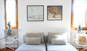 A room with two sofas, a metal chair, a window, and two framed artworks on the wall.