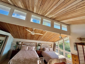 A bedroom with a wooden ceiling, two beds, a ceiling fan, and large windows.
